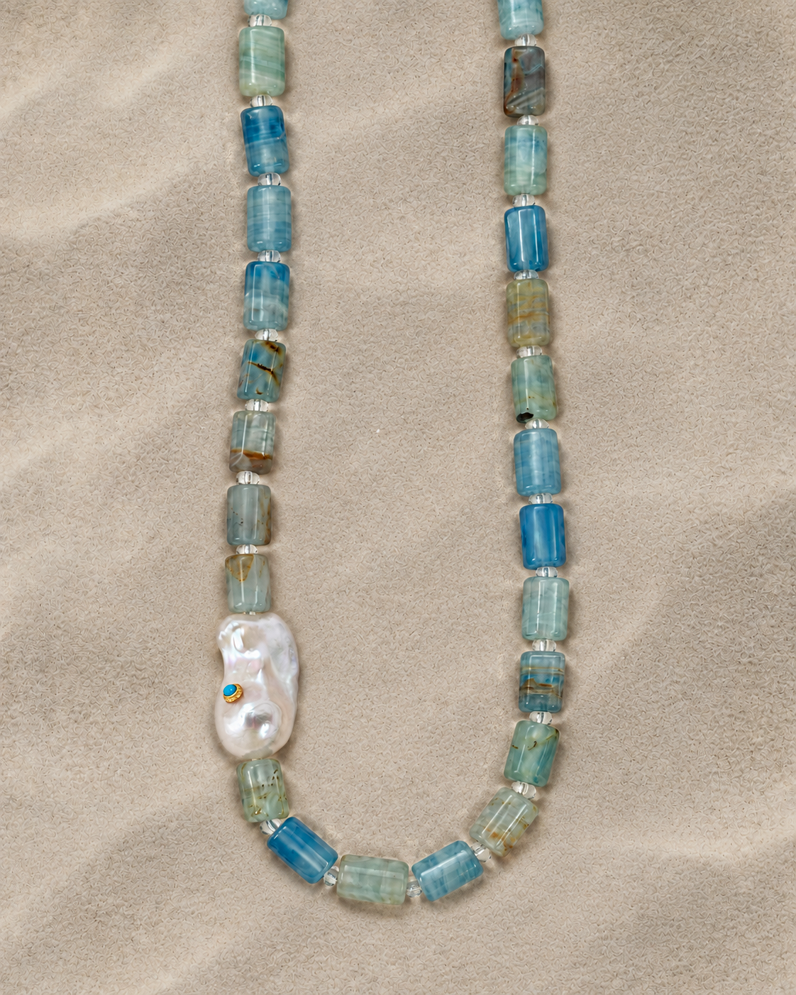 Blue-green stone bead necklace with a sculptural pearl on a warm sand-textured background