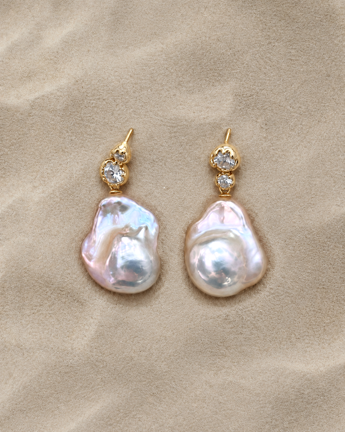 Gold-tone zircon and baroque pearl drop earrings on a sand-textured background